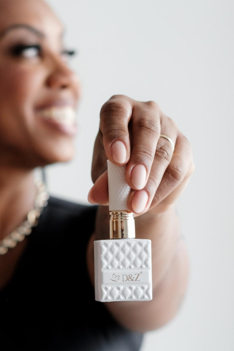 Fabiola com unhas em tom nude segurando um frasco de esmalte; o rosto aparece suavemente desfocado